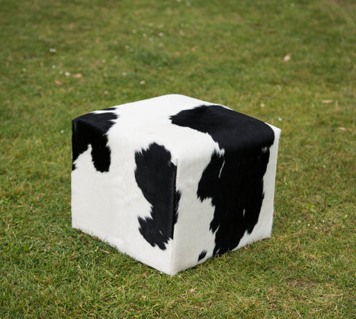 A cube-shaped pouf ottoman with a black and white cow print pattern, placed on a green grass lawn.