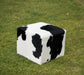 A cube-shaped pouf ottoman with a black and white cow print pattern, placed on a green grass lawn.